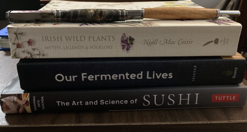 Metal and elderwood fountain pen with magnetic closure next to a seam ripper sheathed in wood. They sit on top of "Irish Wild Plants" by Niall Mac Coitir, "Our Fermented Lives" by Julia Skinner, and "The Art and Science of Sushi" by Mitose Tsuchida.